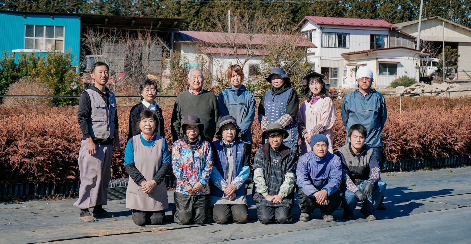 晴れた日の青空の下、二列に並んだ13人の男女。前列には6人がしゃがみ、後列には7人が立っている。彼らの背後には家屋と茶色い低木が見える。参加者は様々な年齢で、仕事中の装いをしている。